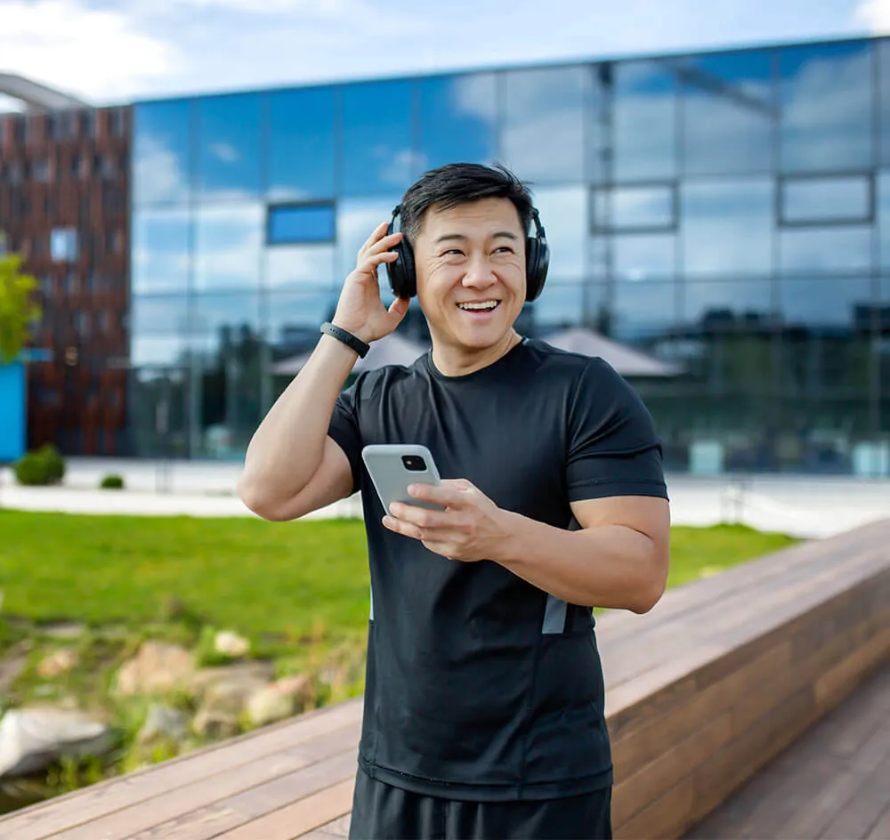 Happy Asian young man doing sports and running near the lake in the city, wearing headphones and holding a phone.