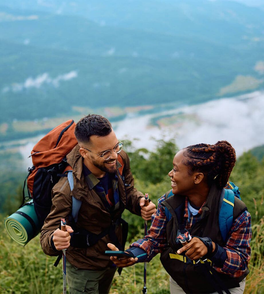 Happy African American hiker and her boyfriend laughing while using cell phone on a hill.