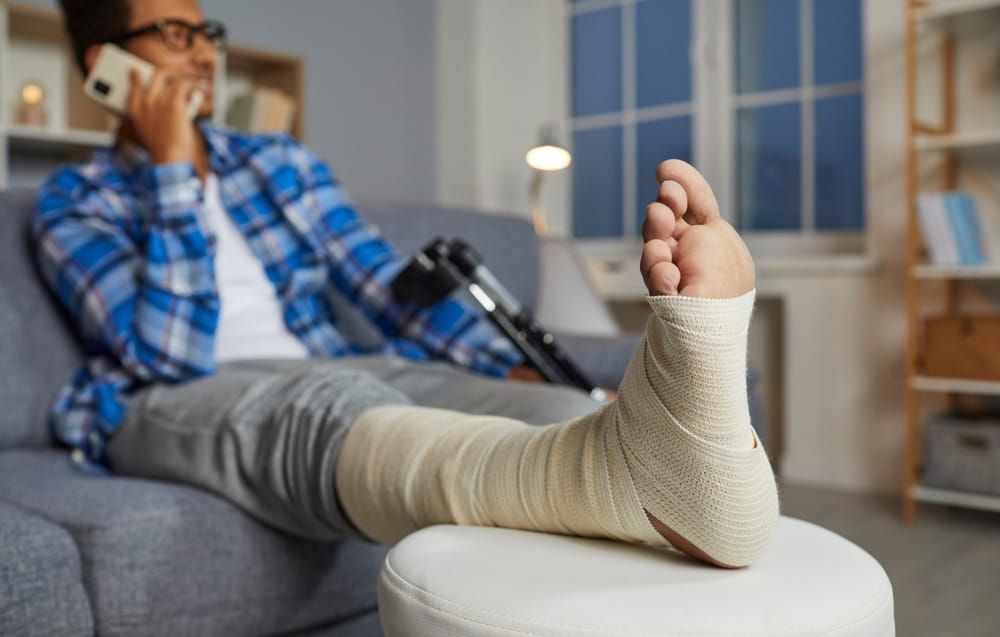 man talking on the mobile phone while relaxing on the sofa at home, with his foot and leg in a bandage resting on a stool