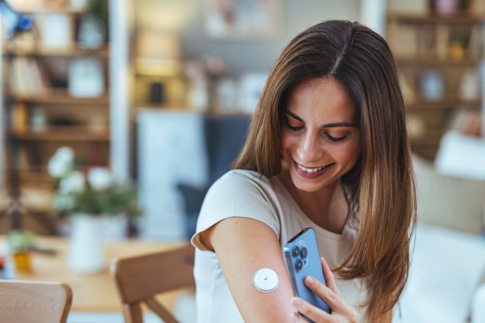 A cheerful woman in a living room checks her glucose levels using a smartphone app connected to a sensor on her arm, illustrating modern diabetes management.