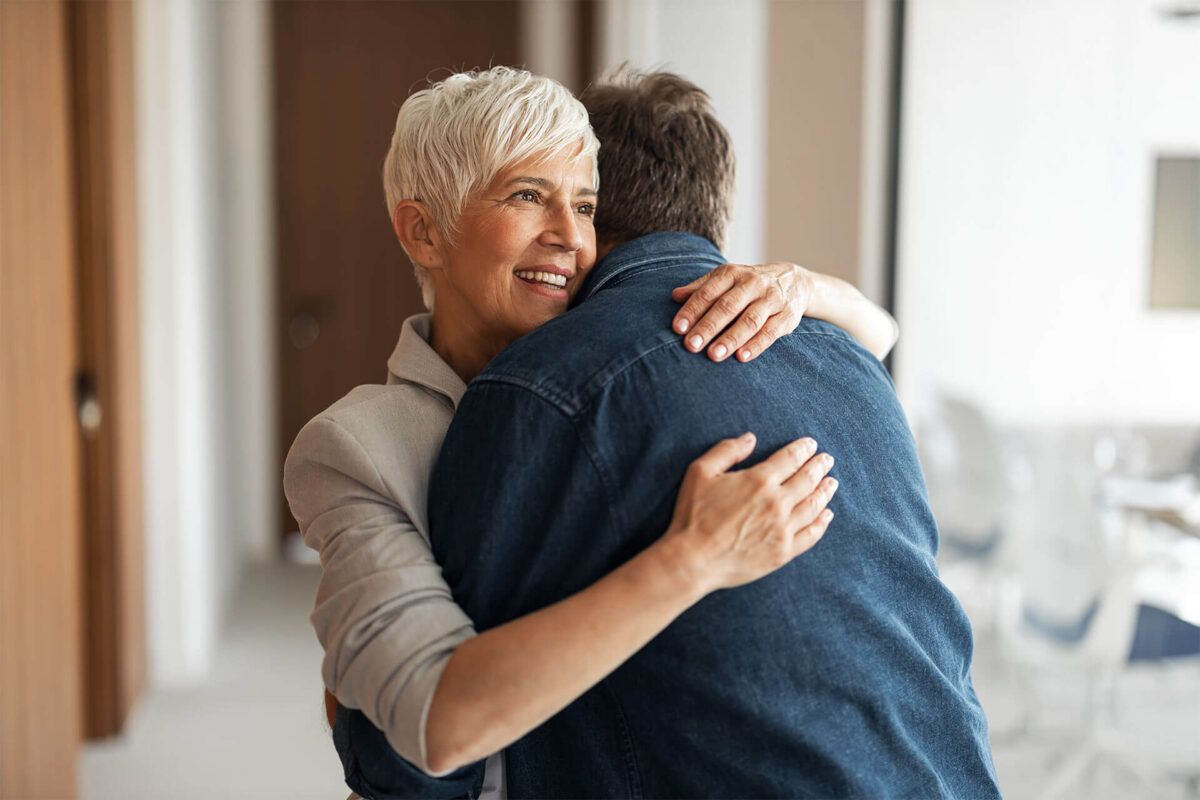 An older woman with short white hair warmly hugs a friend in a denim jacket