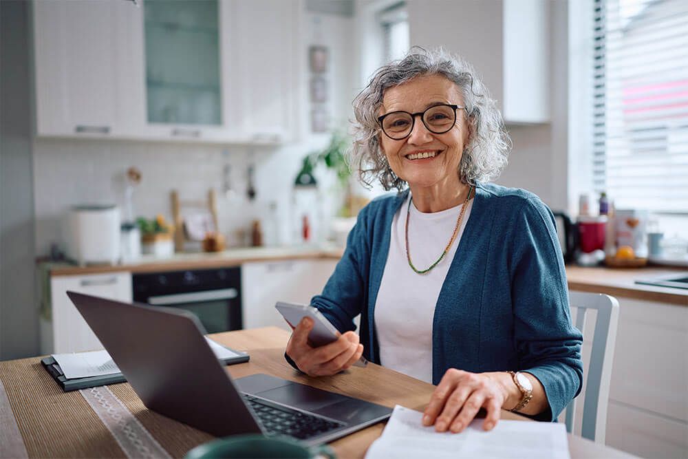 Happy senior woman using cell phone and laptop while going through home budget and looking at camera.