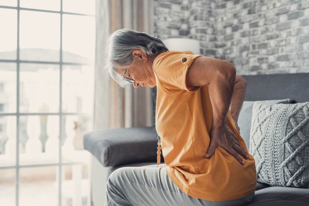 Unhappy mature woman touching back, feeling pain, sitting on couch in living room