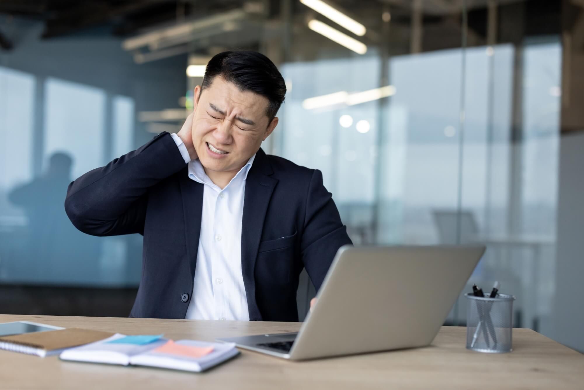 Tired young Asian male businessman sitting in the office at the desk and suffering from neck pain, rubbing it with his hand.