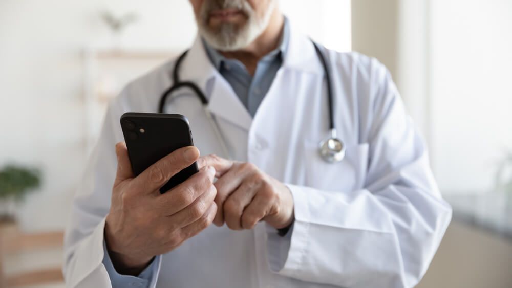 Crop close up of mature male doctor in white uniform hold smartphone browse internet in clinic