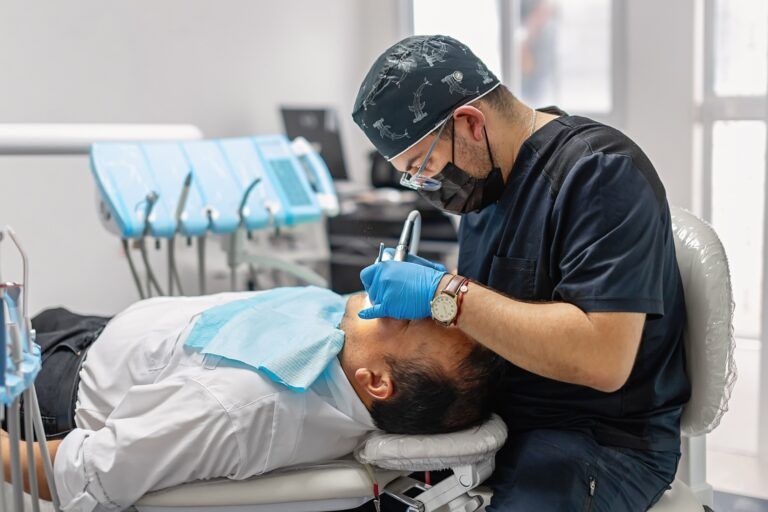 Male dentist working on a patient in a modern dental office. The patient lies relaxed in the chair while the professional conducts a check-up or treatment using gloves and dental tools.