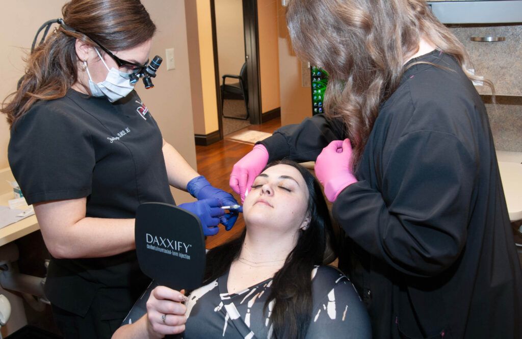 Dr. Yailin Diaz Pichardo applying cosmetic injection procedure, likely Daxxify Botox, to a patient sitting on a chair, with an assistant