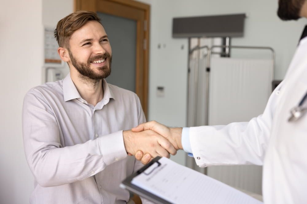 Cheerful positive young Caucasian male patient giving handshake to doctor in lab coat, thanking physician man for medical advice, help, consultation, smiling, shaking hands with medical practitioner